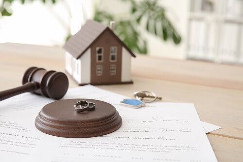 A gavel, two rings, a mini house, keys and property division paperwork on a table.